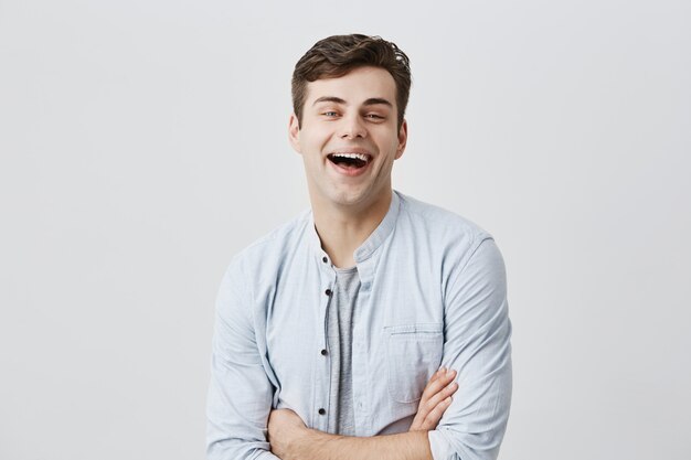陽気な若いヨーロッパ人は歯を広く笑って、肯定的なニュースや仕事での昇進を喜び、腕を組んでいます。人間の感情、感情、態度、反応