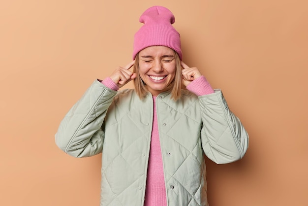 Free photo cheerful young european woman plugs ears keeps eyes closed smiles toothily wears jacket and pink hat avoid loud sound doesnt want to hear you isolated over beige background ignores conversation