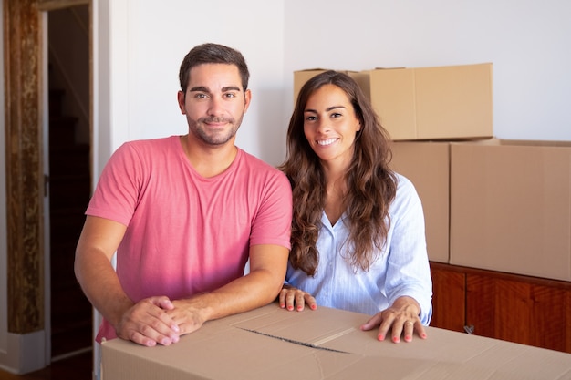 Cheerful young family couple moving into new house, standing at carton box, 