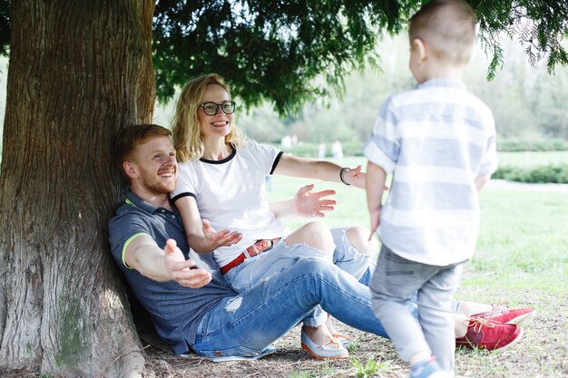 Cheerful young family of mom, dad and little son have fun playing under the green tree