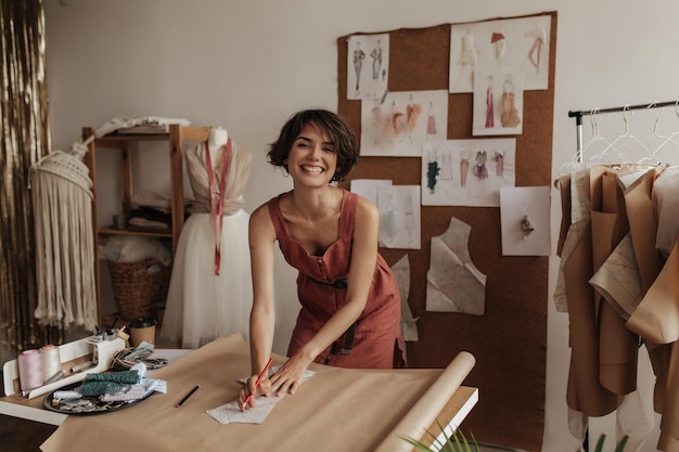 Cheerful young fashion designer in linen red dress smiles sincerely Charming woman holds pencils and draws cloth pattern on craft paper sheet