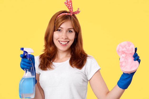 Веселая молодая домработница носит повседневную футболку и повязку на голову, держит моющий спрей и губку, собирается чистить пыль