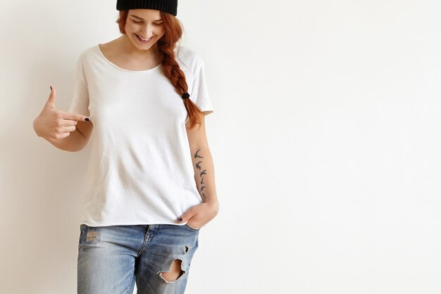 Cheerful young female with ginger hair and tattoo looking down and pointing index finger at her white oversize t-shirt
