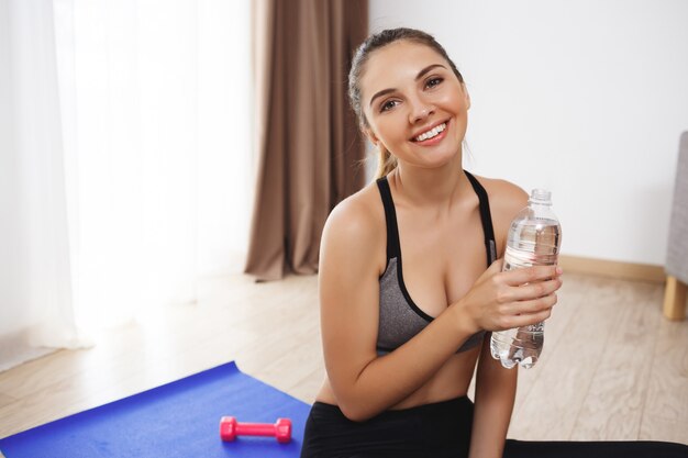 Cheerful young fitness girl make yoga exercises on floor and drink water