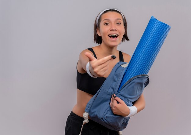 Веселая молодая фитнес-девушка в спортивной одежде держит рюкзак с ковриком для йоги, счастливым и позитивным, делая жест, стоящий над белой стеной