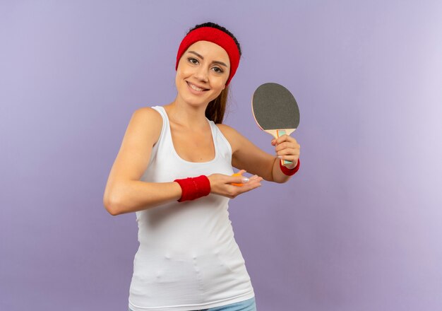 Веселая молодая фитнес-девушка в спортивной одежде с повязкой на голову держит ракетку и мяч для настольного тенниса с улыбкой на лице, стоящей над серой стеной