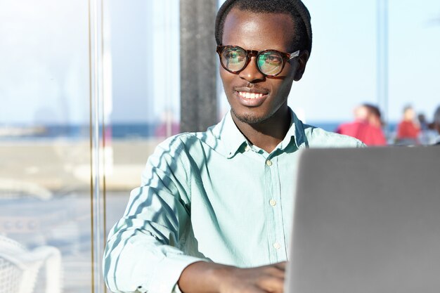 Cheerful young freelancer in a trendy hat and eyewear smiling happily