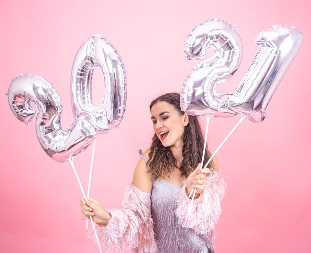 Cheerful young girl in festive outfit on pink studio background posing holding silver balloons for the new year concept