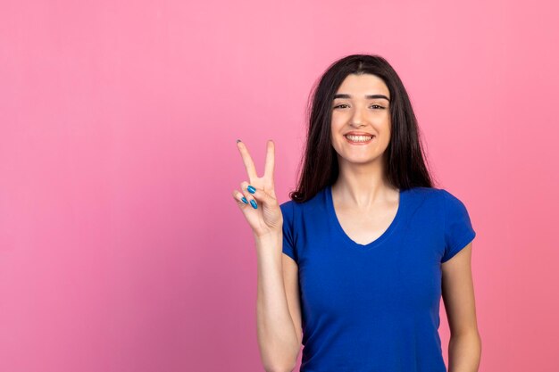 Веселая молодая девушка жест мира и улыбка на розовом фоне Фото высокого качества