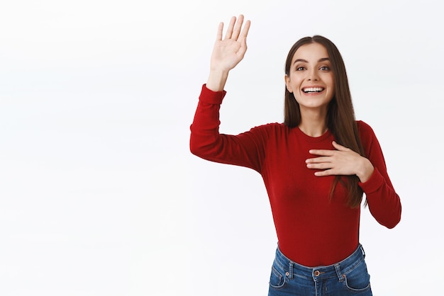 Cheerful young girl see friend in crowd trying attract attention as waving raised hand in hello or hi gesture say over here as smiling and jumping to be seen standing white background joyful