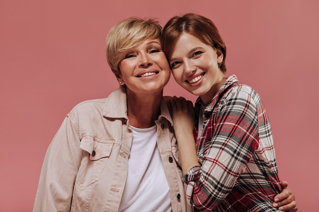 Cheerful young girl with short hairstyle in plaid shirt looking into camera and smiling together with blonde woman in beige jacket on pink backdrop. 