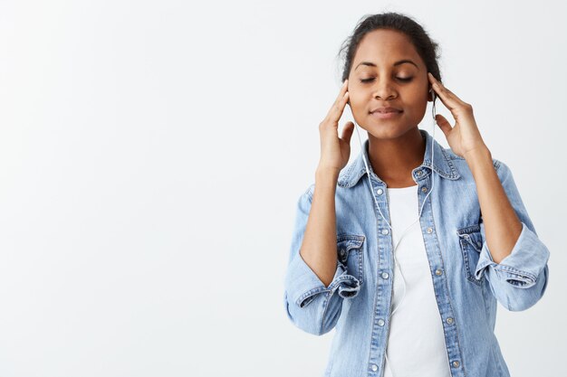 Cheerful young good looking woman with clean dark skin and black hair posing indoors with earphones on, enjoying her favourite song with closed eyes, wearing casual blue shirt.