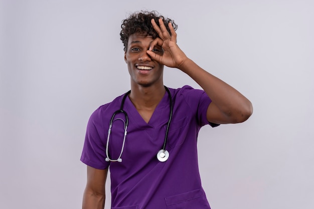 A cheerful young handsome dark-skinned doctor with curly hair wearing violet uniform with stethoscope peeking through a hole formed with his thumb and index finger 