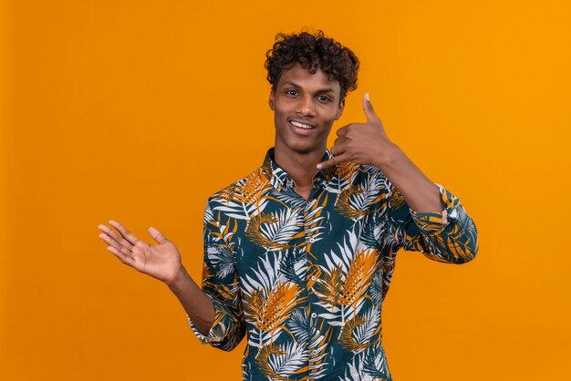 Cheerful young handsome dark-skinned man with curly hair in leaves printed shirt making a call me sign with his hand 