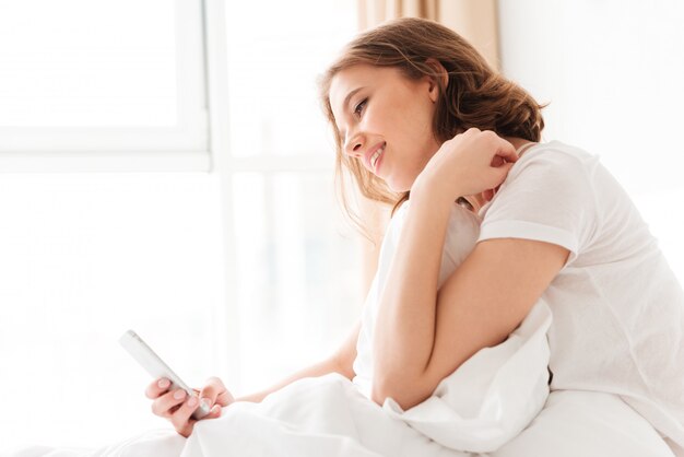 Cheerful young lady in bed indoors looking aside chatting