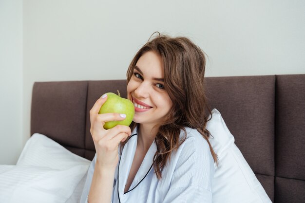 Cheerful young lady dressed in pajama eating apple.