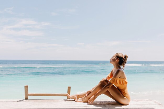 夏の朝のビーチに座っている陽気な若い女性。ビーチでポーズをとってオレンジ色の水着でゴージャスな女の子の屋外ショット