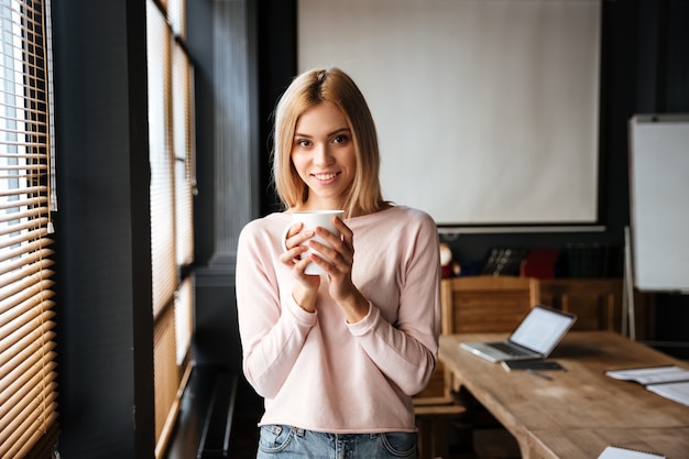 Cheerful young lady standing in cafe drinking coffee