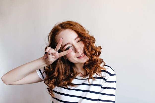 Веселая юная леди с бледной кожей и рыжими волосами позирует со знаком мира. Удивительная девушка в полосатой изолированной футболке.