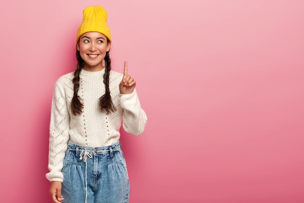 Cheerful young lady with two plaits, raises finger above, promots top copy space against pink background