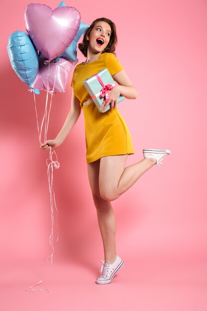 Cheerful young lady in yellow dress holding ballons with gift