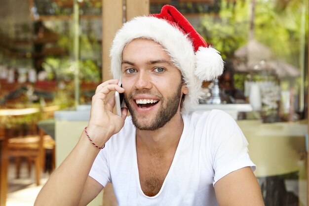 Cheerful young male wearing red Santa Claus hat smiling happily while having phone conversation