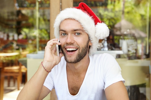 Веселый молодой мужчина в красной шляпе Санта-Клауса счастливо улыбается во время телефонного разговора
