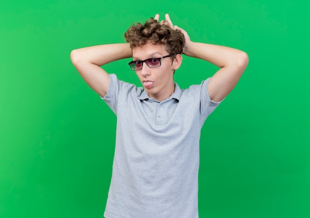 Free photo cheerful young man in black glasses wearing grey polo shirt imitating bunny ears sticking out tongue standing over green wall