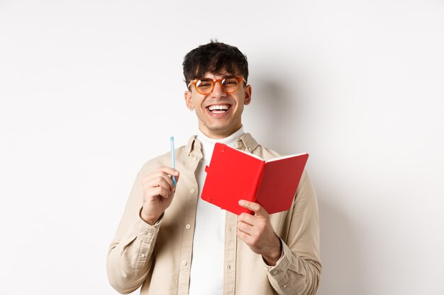 Cheerful young man in glasses laughing and taking notes, writing down in planner, holding pen and diary, standing on white background.