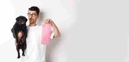 Free photo cheerful young man holding black pug and pink dog poop bag standing over white background