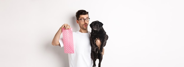 Cheerful young man holding black pug and pink dog poop bag standing over white background