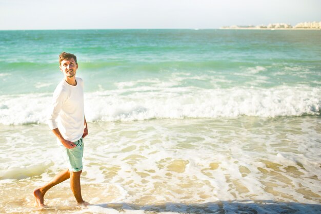 Cheerful young man looks up in the sky walking in foaming waves