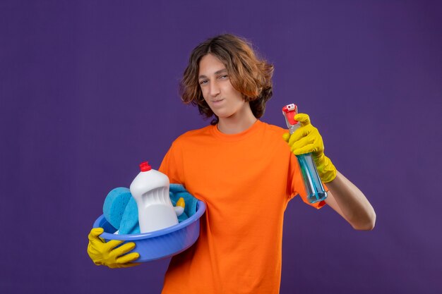 Веселый молодой человек в оранжевой футболке в резиновых перчатках держит таз с чистящими средствами и чистящим спреем, положительно и счастливо стоит над фиолетовым пространством