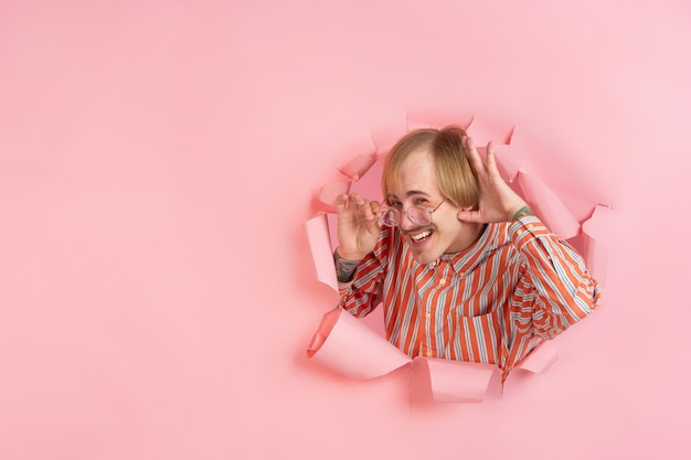 Cheerful young man poses in torn coral paper hole wall emotional and expressive