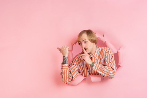 Free photo cheerful young man poses in torn coral paper hole wall emotional and expressive