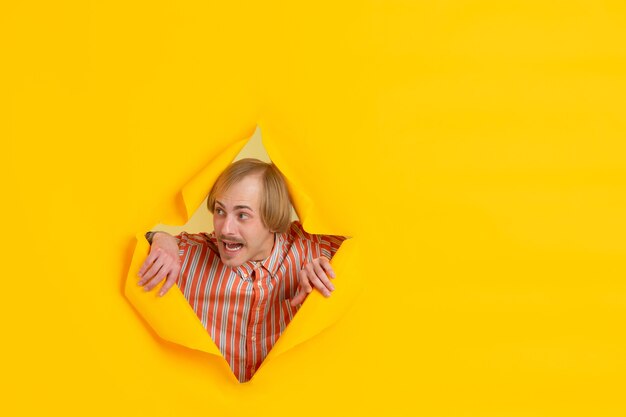 Cheerful young man poses in torn yellow paper hole wall emotional and expressive