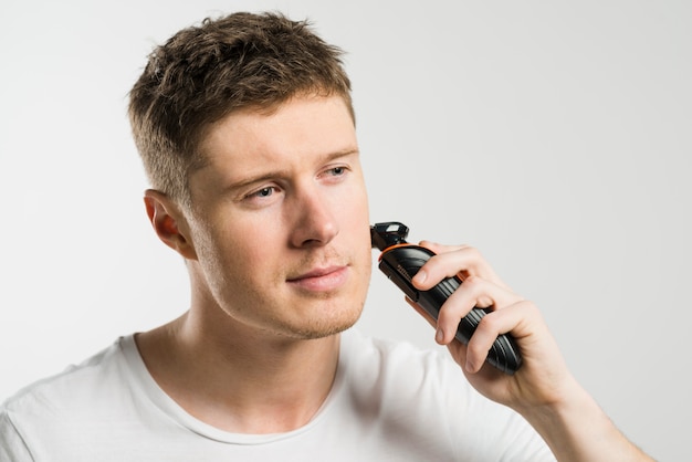 Cheerful young man shaving with machine against white background