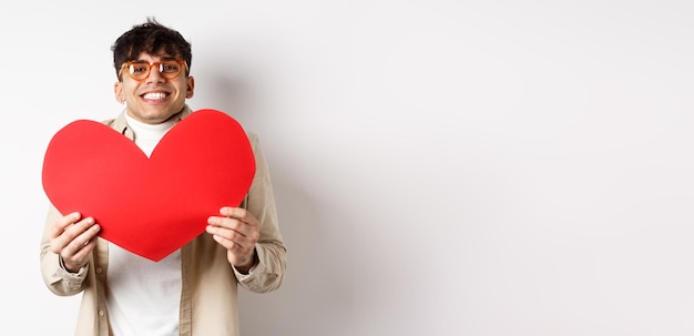 Free photo cheerful young man smiling and looking thrilled at camera showing big red heart cutout on valentines