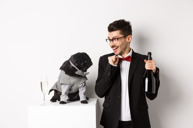 Cheerful young man in suit celebrating New Year with pet, dog and guy looking at each other, owner drinking champagne, standing over white background.