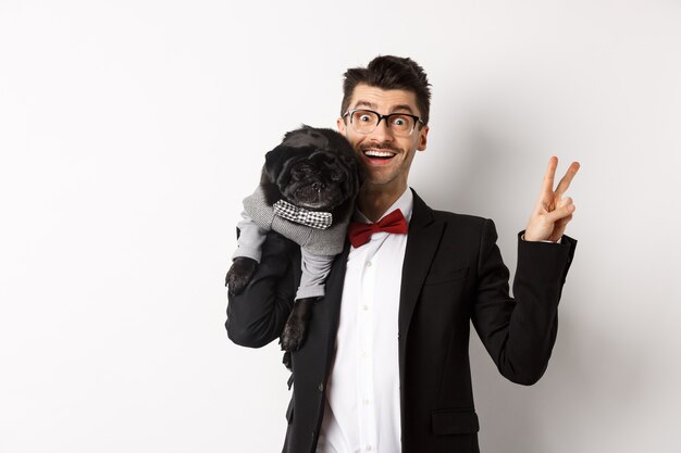 Веселый молодой человек в костюме и очках фотографирует с милой черной собакой мопса на плече, улыбается счастливым и показывает знак мира, позирует на белом фоне.