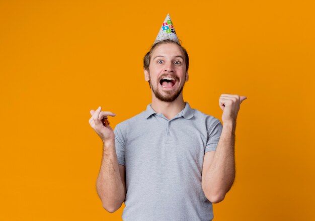 Cheerful young man with holiday cap celebrating birthday party crazy happy and excited standing over orange wall