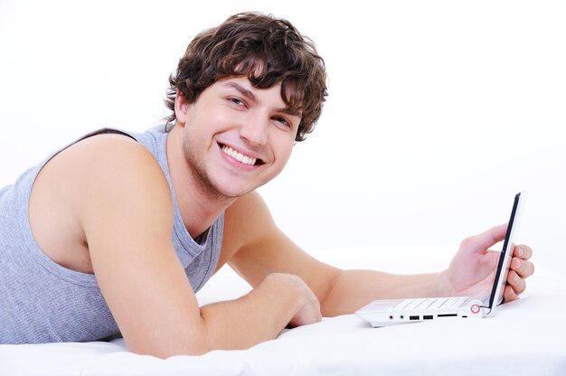 Веселый молодой человек с ноутбуком, лежа в спальне, глядя в камеру