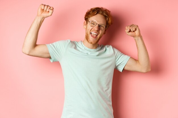 幸せそうに見える赤い髪の陽気な若い男、拳ポンプジェスチャーで手を上げ、成功を祝って、チャンピオンのように感じ、勝利し、ピンクの背景の上に立っています