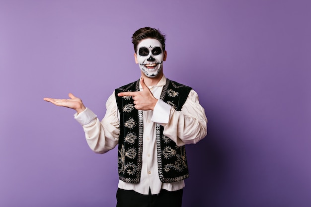 Cheerful young man with sincere smile is pointing finger at his hand. Indoor snapshot of guy with Halloween makeup with space for text on isolated wall.