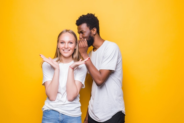 Cheerful young mixed couple whispering secret behind her hand sharing news posing isolated on yellow wall