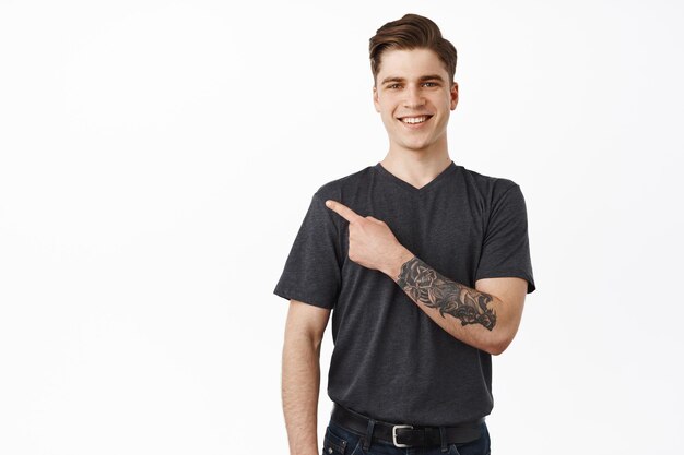 Cheerful young modern man, handsome guy with tattoo smiles, pointing left and showing logo banner on white empty space, standing against studio background.