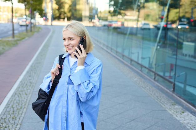 Cheerful young modern woman walking on street and answers phone call talking on mobile carries black