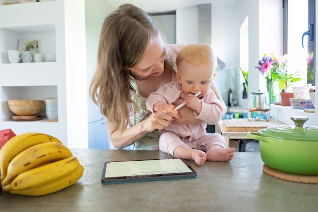 쾌활 한 젊은 엄마와 함께 집에서 요리, 태블릿을 사용 하여 패드에 조리법을보고 아기 딸. 육아 또는 가정에서 요리 개념