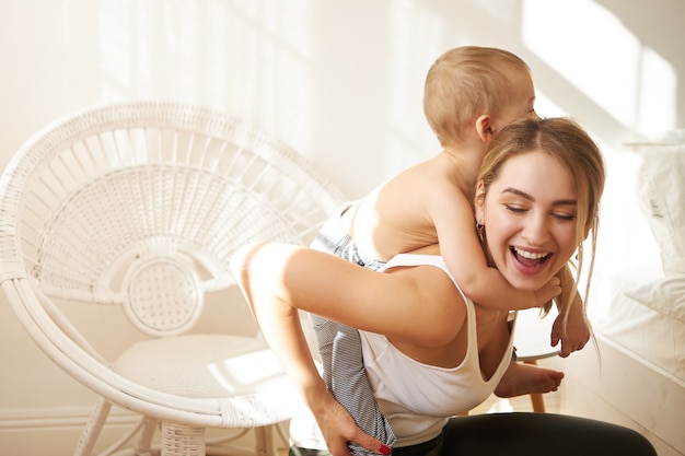 Cheerful young mother having fun indoors giving piggy back ride to her cute toddler son. Attractive mom spending time in nursery, playing with child during social distancing because of quarantine