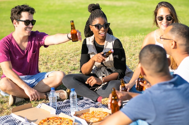 Cheerful young people cheering with beer bottles in park. Happy friends sitting on meadow and drinking beer. Leisure concept